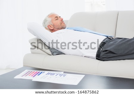 Side view of a tired mature businessman sleeping on sofa with graphs on table in the living room at home