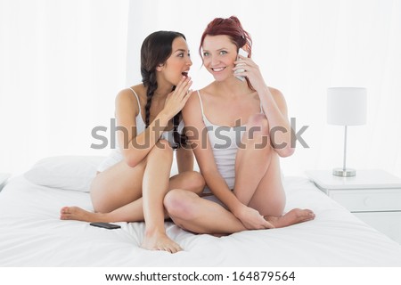 Full length of two happy young female friends using mobile phone while sitting on bed at home