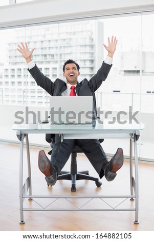 Elegant businessman cheering with raised hands in front of laptop at office desk