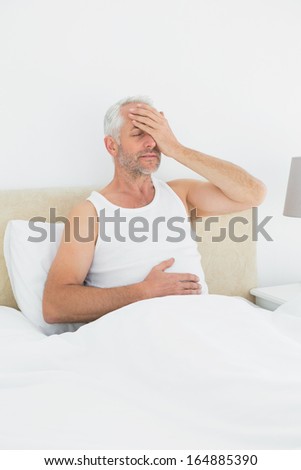 Thoughtful mature man sitting in bed with hand over closed eyes at home