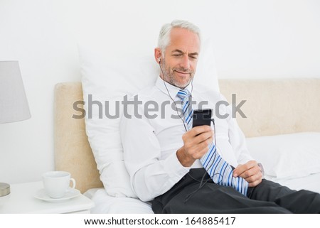 Smiling well dressed man listening music with his phone in bed at home