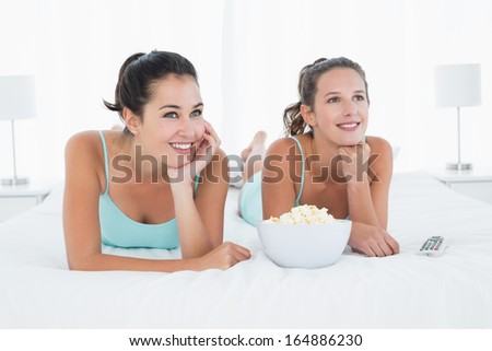 Two smiling young female friends with popcorn bowl lying in bed at home