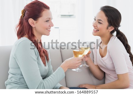 Side view of two happy young female friends toasting wine glasses on sofa at home