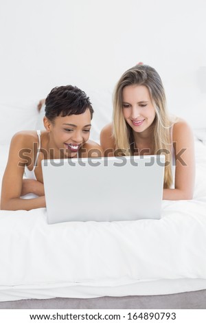 Happy relaxed young female friends using laptop in bed at home