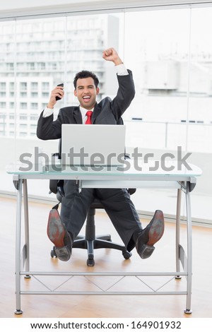 Elegant businessman cheering with clenched fists in front of laptop at office desk
