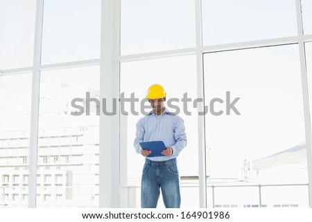 Smiling young architect in yellow hard hat with clipboard in a bright office