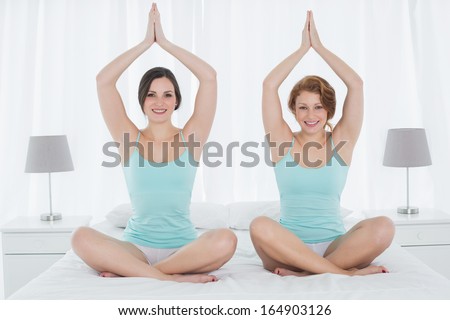 Full length portrait of two young women sitting with joined hands over head on bed at home