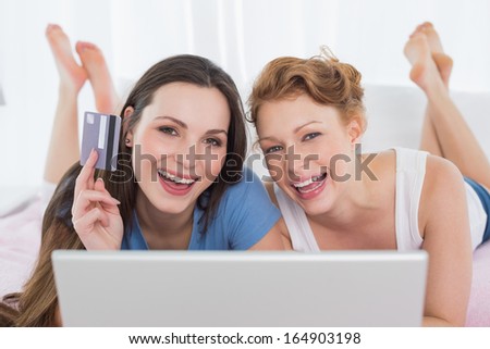Portrait of cheerful relaxed young female friends  doing online shopping through laptop and credit card in bed
