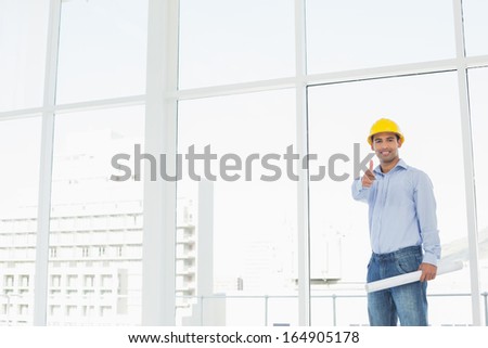 Portrait of a smiling architect in yellow hard hat with blueprint gesturing thumbs up in a bright office