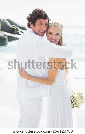 Smiling bride and groom looking at camera hugging at the beach