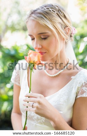 Smiling blonde bride in pearl necklace smelling rose in the countryside