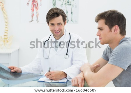 Smiling male doctor writing reports besides patient in the medical office