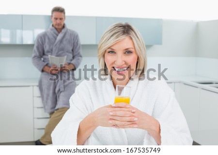 Woman with orange juice while man reading newspaper in background at the kitchen