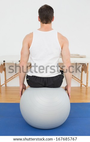 Rear view of a young man sitting on yoga ball in the gym at hospital