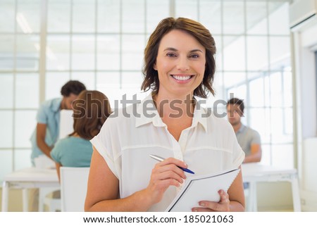 Portrait of a smiling businesswoman writing notes up with colleagues in meeting in background at the office