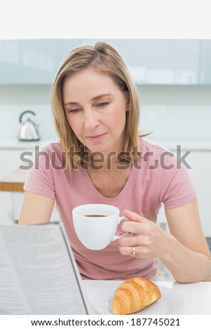 Concentrated woman reading newspaper while having coffee in the kitchen at home