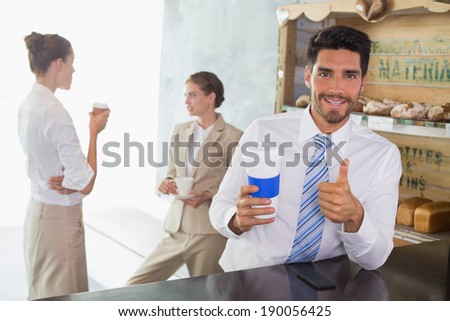 Businessman with coffee sipper gesturing thumbs up at the counter with colleagues behind in office cafeteria
