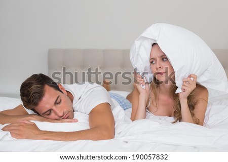 Angry young woman holding pillow besides a sleeping man in bed at home