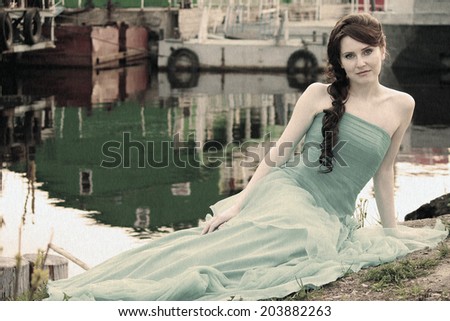 Beautiful brunette girl in blue wedding dress sitting on grass at riverbank at sunset. Image with noise vintage filter