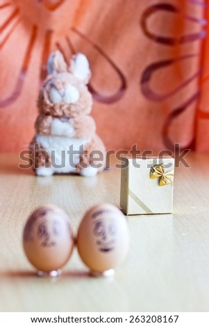 Painted easter eggs with man and woman smiling faces. Eggs on wedding rings. Blurred background with golden gift box and easter bunny. Conceptual funny image. Focus on gift box