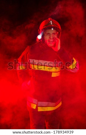 Shouting fireman in red helmet holding axe and standing in smoke. Three quarter length studio shot on black background.
