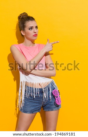 Beautiful young woman pointing and looking away. Three quarter length studio shot on yellow background.