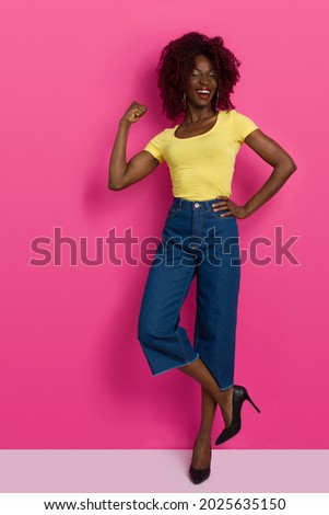 Confident and happy young black woman in yellow top, jeans and high heels is standing on one leg, flexing muscles and laughing. Full length studio shot on pink background. Foto stock © 