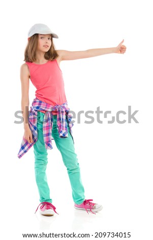 Young girl in baseball cap and green trousers showing thumb up. Full length studio shot isolated on white.