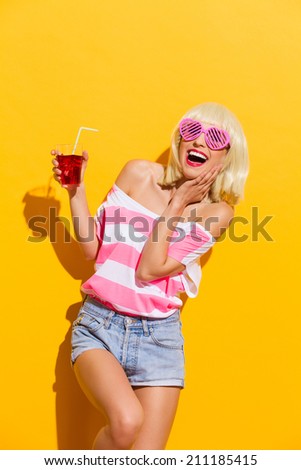 I love this cold drink. Blond young woman in pink sunglasses holding red drink and laughing. Three quarter length studio shot on yellow background.