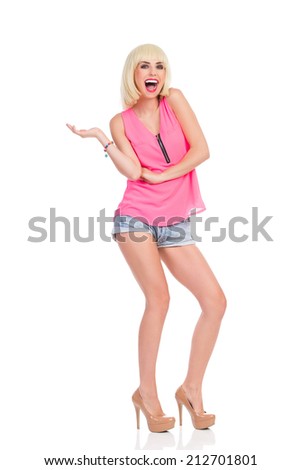 Shouting blond young woman in high heels and pink top standing and presenting something on her hand. Full length studio shot isolated on white.