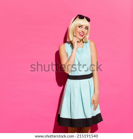 Sideways glance. Smiling young woman standing in a sunlight against pink wall, holding hand on chin and looking away. Three quarter length studio shot on pink background.