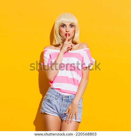 It\'s a secret. Beautiful blonde young woman holding finger on her lips. Three quarter length studio shot on yellow background.