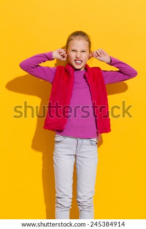 I don\'t want to hear it. Young blond girl stops her ears. Three quarter length studio shot on yellow background.