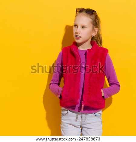 Cheerful blond girl in pink fur vest posing with hands in pockets. Waist up studio shot on yellow background.