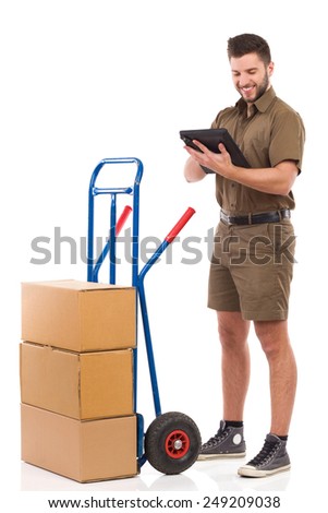 Messenger working on a digital tablet. Smiling courier standing close to push cart and using a digital tablet. Full length studio shot isolated on white.