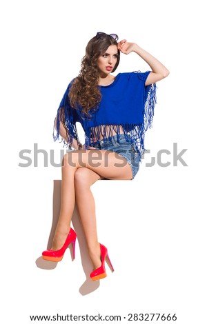 Sexy young woman in blue top, jeans shorts and red high heels sitting on a top of the white banner with legs crossed at knee and looking away. Full length studio shot isolated on white.