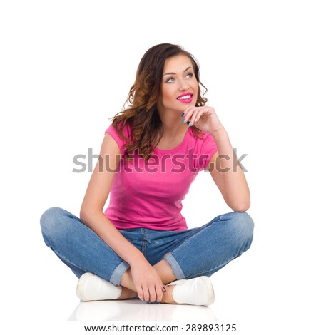 Sitting Female Student Looking Away And Thinking. Cheerful young woman in pink shirt and jeans sitting on a floor with legs crossed and looking away. Full length studio shot isolated on white.