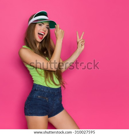 Peace Woman! Shouting young woman in lime green shirt and green sun visor showing a peace sign. Three quarter length studio shot on pink background.