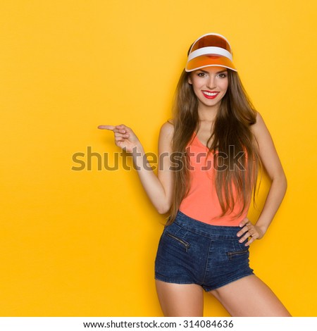 Hey! Look here. Cheerful young woman in orange shirt and sun visor pointing at copy space. Three quarter length studio shot on yellow background.