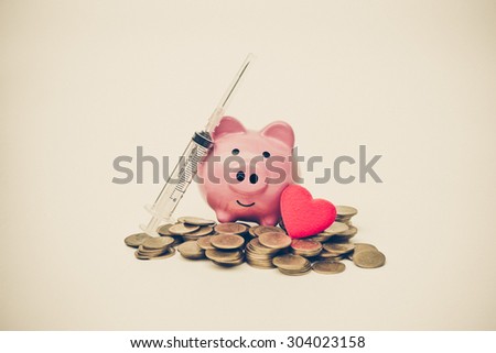 a pink piggy bank, a syringe with needle, and a red heart on piles of golden coins - healthcare cost concept
