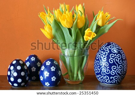 Fresh yellow tulips blossoming in a glass vase and blue ceramic easter eggs in front of warm earthy colored wall