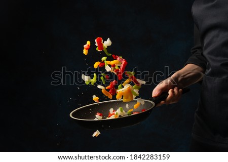 Close-up view of chef's hand throws up frying mix of colored vegetables above the pan on dark blue background. Backstage of cooking meal. Frozen motion. Food banner concept.