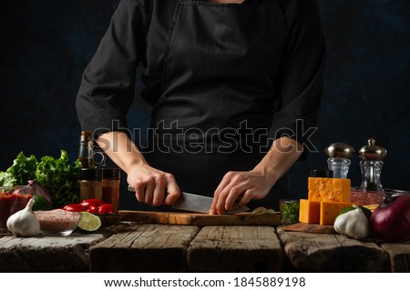 Professional chef in black uniform cuts with knife garlic on chopped wooden board. Backstage of cooking traditional mexican tacos on rustic wooden table. Close-up view. Dark blue background.
