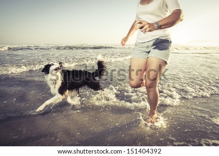 Young woman running with her dog in the shallow water on the beach.