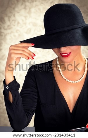 Attractive woman wearing black dress, hat and pearls, sitting on black table.