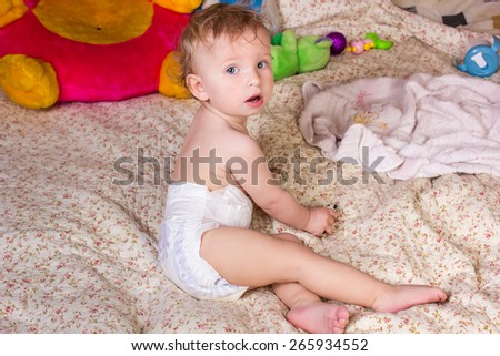 Cute blonde baby girl with beautiful blue eyes sits on bed in diapers with toy