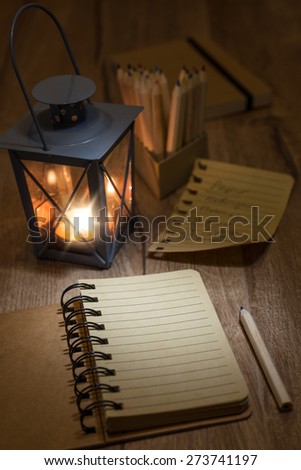 Open notebook with blank front page, candle in metal vintage lantern, torn-out page and a pencil on wooden table. Space for your notes, recipe or a poem on the page.