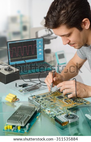 Young energetic male tech or engineer repairs electronic equipment in research facility. Shallow DOF, focus on the face of the worker.