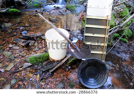 Basic tools used for placer gold prospecting. These include a gold pan, sluice box, 5 gallon plastic bucket and a shovel. A peaceful mountain stream is used for a backdrop.
