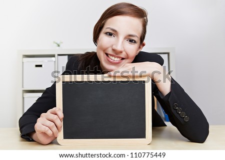 Smiling female teacher sitting with empty black chalkboard at her desk
